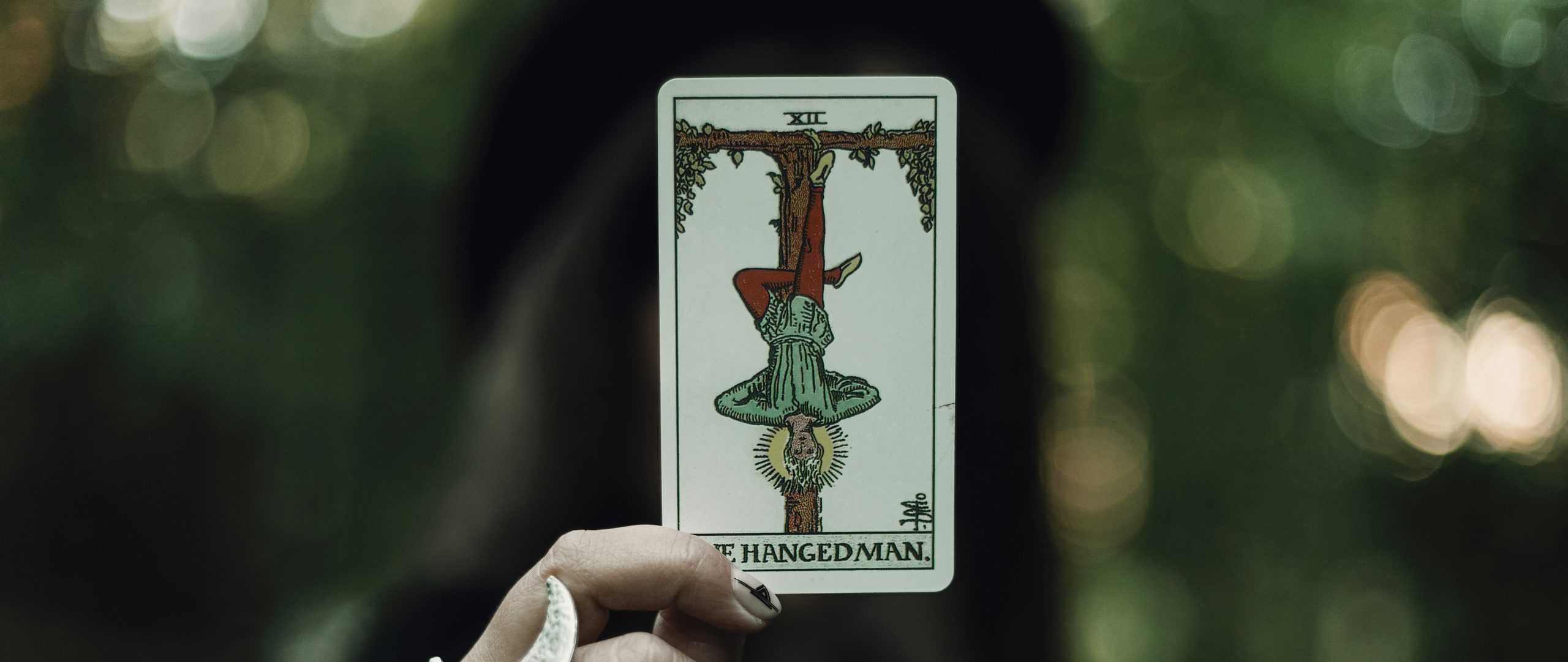 La carta XII del Tarot, El Colgado, sostenida en alto frente a un bosque desenfocado